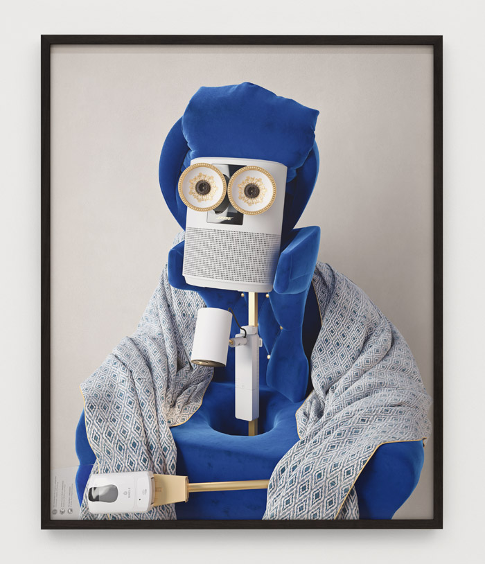 A wood-framed portrait of an androgynous figure in a regal and ornate costume made of shiny material. The figure is standing against a dark background and is holding a staff. The overall scene evokes a futuristic and otherworldly aesthetic.